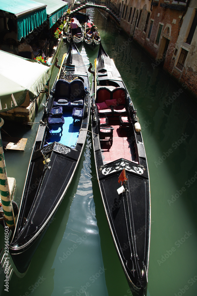 Two black Venetian gondolas with crimson and blue upholstery are on a narrow canal at a cafe