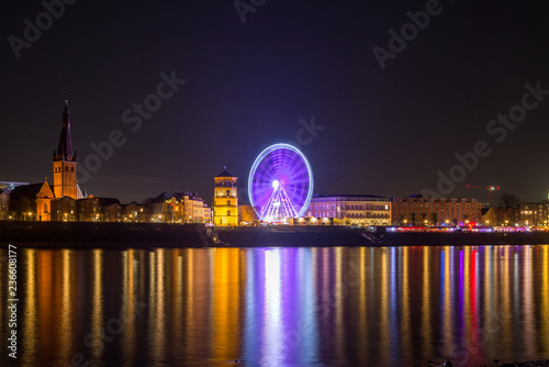 Night scenery colorful illuminated and reflected on river of buildings in old town, big moving Ferris Wheel and Christmas Market on promenade along riverside Rhine River in Düsseldorf, Germany. 