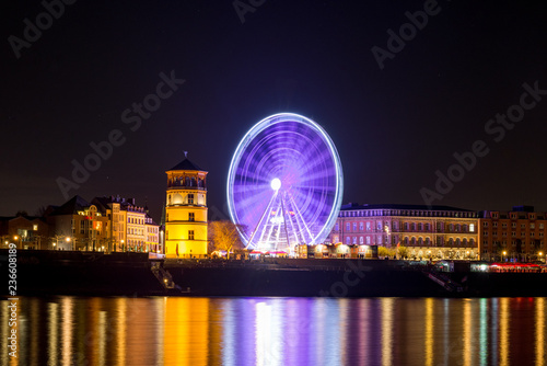 Night scenery colorful illuminated and reflected on river of buildings in old town, big moving Ferris Wheel and Christmas Market on promenade along riverside Rhine River in Düsseldorf, Germany. 