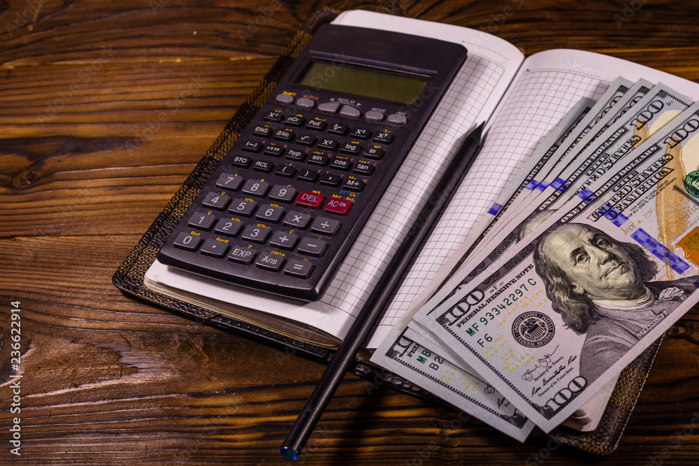 Opened notepad, one hundred dollar banknotes, scientific calculator and pencil on wooden table Opened notepad, one hundred dollar banknotes, scientific calculator and pencil on wooden table