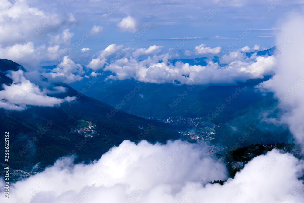 view from above, from the peak or from an airplane, to a wooded mountain valley with settlements, partly hidden by cumulus clouds