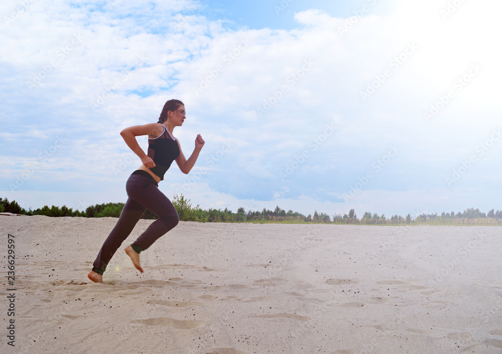 Sporty young girl running through the desert on the sand near the forest sun shining.