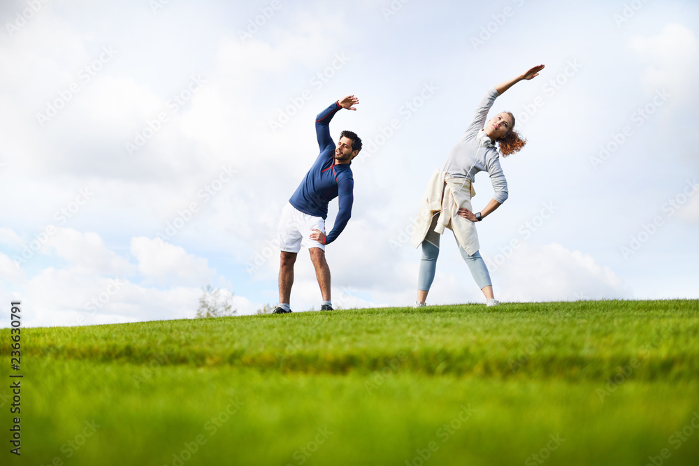 Active guy and girl in sportswear doing side bend exercise while standing on green field Active guy and girl in sportswear doing side bend exercise while standing on green field