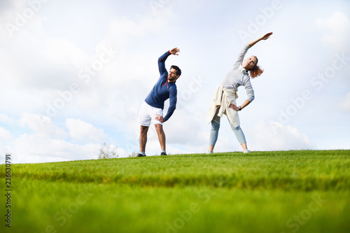 Active guy and girl in sportswear doing side bend exercise while standing on green field