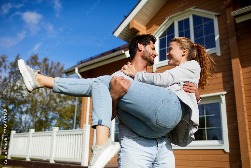 Young man holding his happy wife on hands on background of their new house