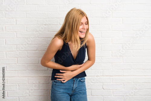 Blonde woman smiling a lot while putting hands on chest on white brick wall background