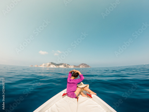 Young woman sitting on bow of boat in the middle of the sea. Happy girl with hat raising hands enjoying freedom travel. © WineDonuts