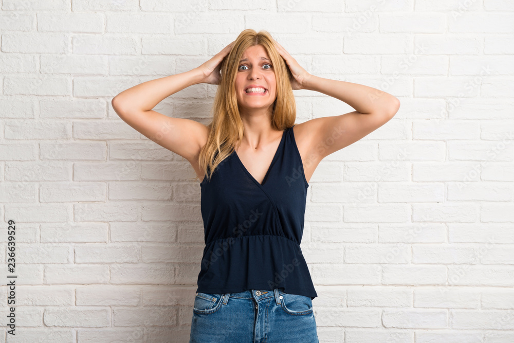 Blonde woman takes hands on head because has migraine on white brick wall background