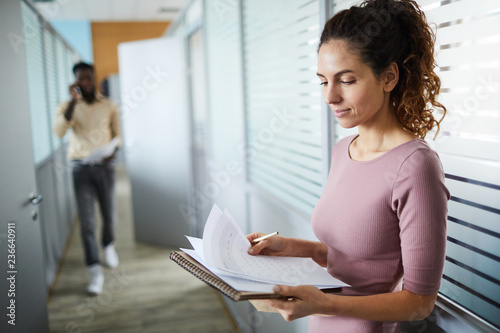 Young businesswoman standing in corridor by office and looking through financial papers