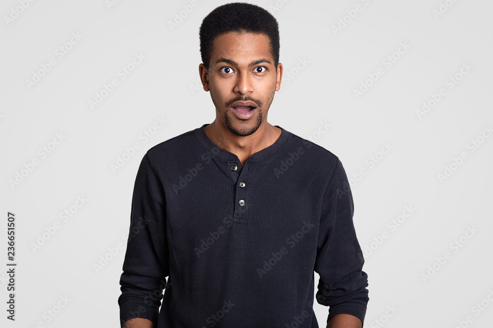 Indignant stupefied dark skinned young guy opens mouth with surprisement, dressed in casual jumper, reacts on sudden news from interlocutor, isolated over white background. Masculinity concept Indignant stupefied dark skinned young guy opens mouth with surprisement, dressed in casual jumper, reacts on sudden news from interlocutor, isolated over white background. Masculinity concept