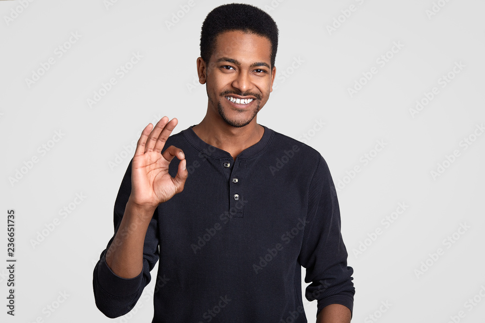 Everything is all right or great! Positive smiling hipster guy with dark healthy skin, shows okay gesture, approves good idea of friend, stands against white background. Body language concept Everything is all right or great! Positive smiling hipster guy with dark healthy skin, shows okay gesture, approves good idea of friend, stands against white background. Body language concept