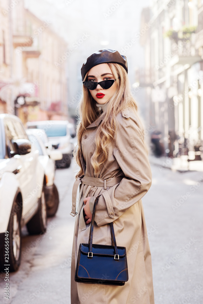Obraz premium Outdoor fashion portrait of young beautiful fashionable girl wearing trendy beige long trench coat, stylish cat eye sunglasses, leather beret, holding handbag, model posing in street of european city