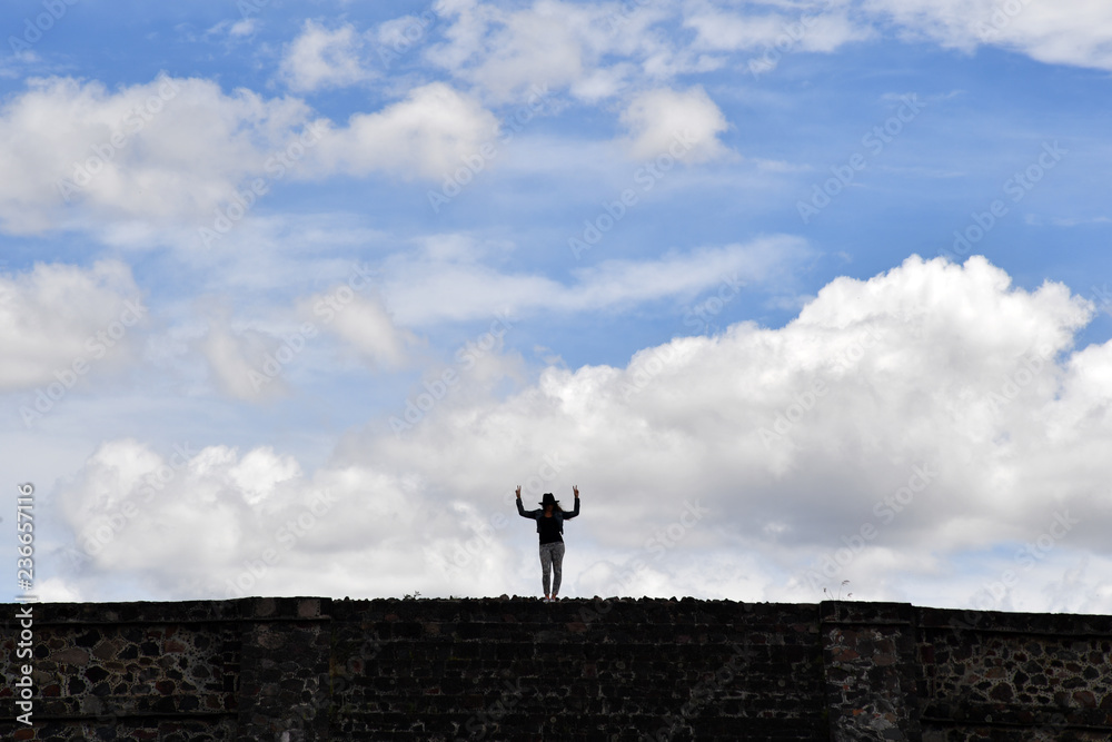 A happy young woman on top of the sun pyramid in Teotihuacán raises her hands in the sky. 