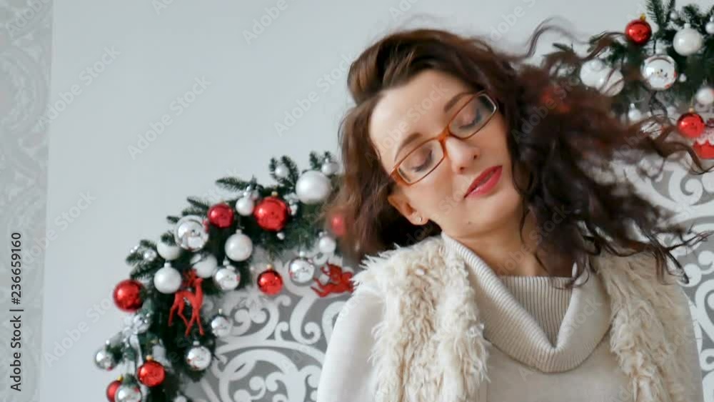 Portrait of a happy brunette girl wearing eyeglasses and warm sweater on the background of Christmas decor. Curly girl laughs Portrait of a happy brunette girl wearing eyeglasses and warm sweater on the background of Christmas decor. Curly girl laughs