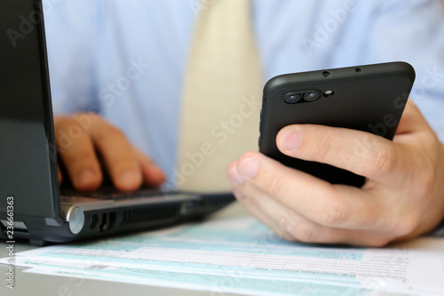 Businessman in tie works in office, sitting at a table with laptop and smartphone in hand. Male hands close-up, concept of clerk, official or bank employee using notebook, typing on keyboard