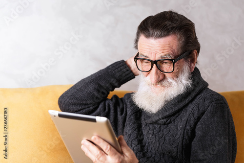 Senior bearded man in glasses wearing grey pullover sitting on a yellow sofa in his light living room and looking doubtfully on the digital tablet photo