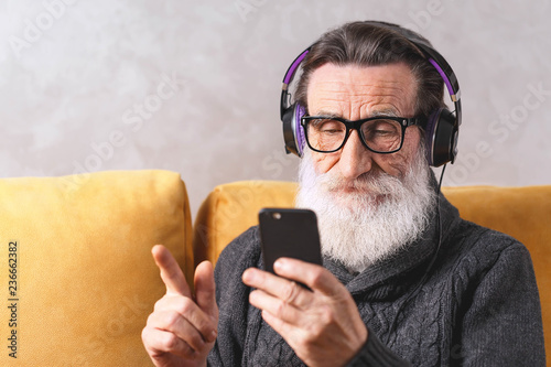 Senior contemporary bearded man in glasses wearing grey pullover learning to use the smartphone while sitting in headphones on a yellow sofa in his light living room, modern technology, communication