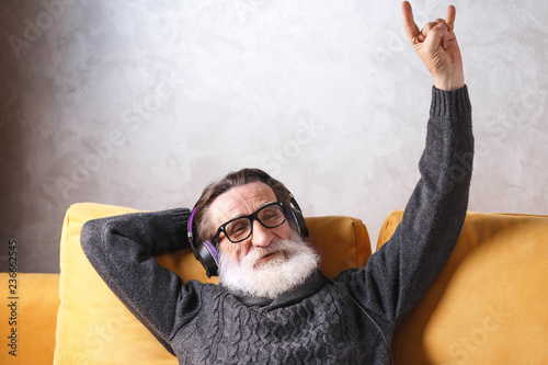 Senior handsome bearded man in glasses wearing grey pullover sitting in headphones on a yellow sofa in his light living room, relaxing and listening his favorite rock music song, modern technology