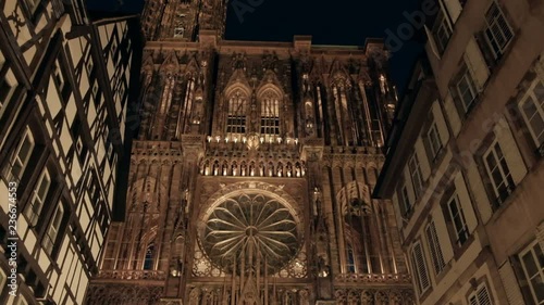 Reverse push-out camera tilt up from base to tower of lighted Notre Dame Cathedral in Petite France old town at night between buildings in Strasbourg, France