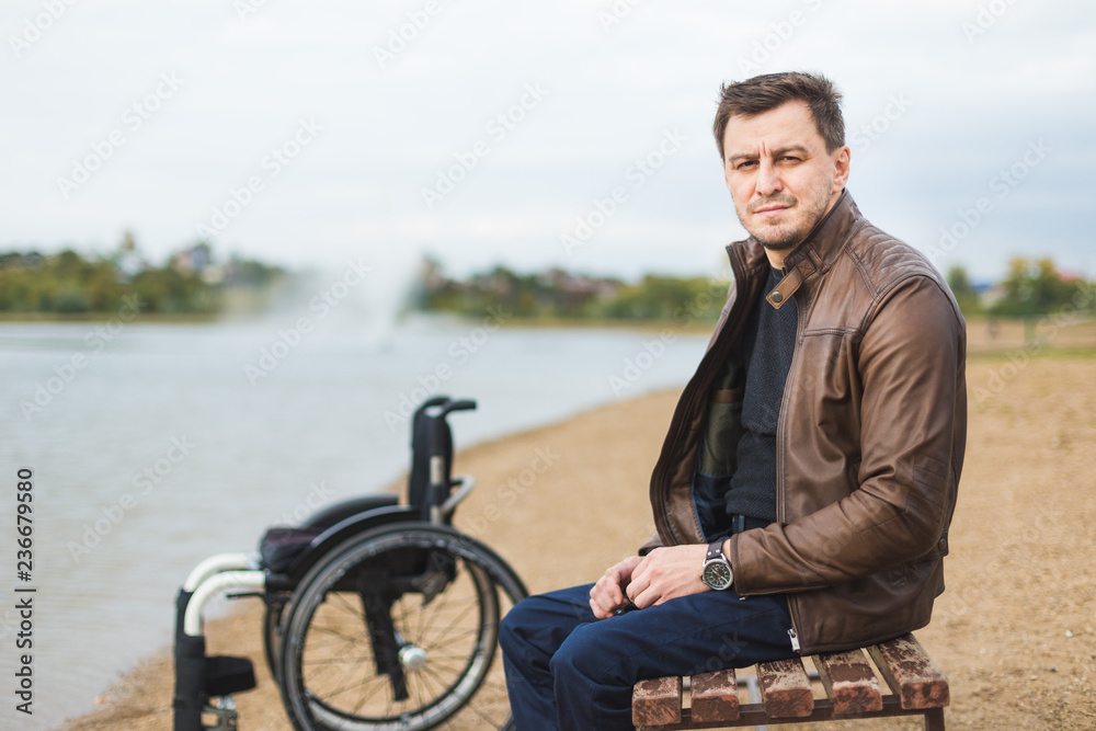 A young man sits on a bench by the lake, next to his wheelchair.