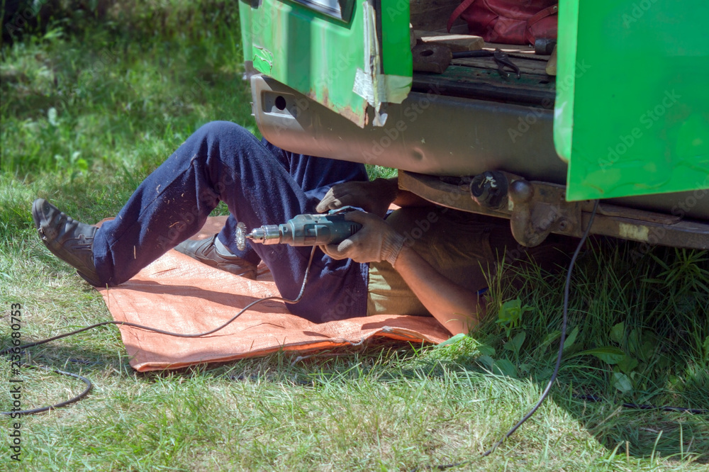The mechanic lies under the car and repairs the body of a green minibus in the summer on the green grass outside. The mechanic lies under the car and repairs the body of a green minibus in the summer on the green grass outside.