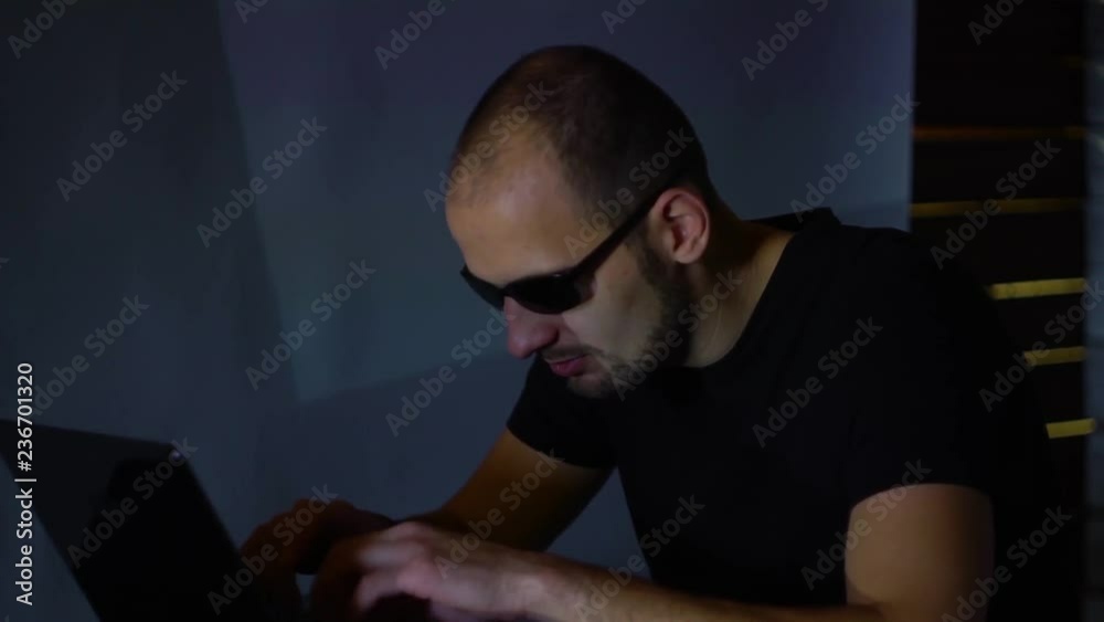 A man hacker in sunglasses in a dark room works with the program code. concept on cybercrime A man hacker in sunglasses in a dark room works with the program code. concept on cybercrime