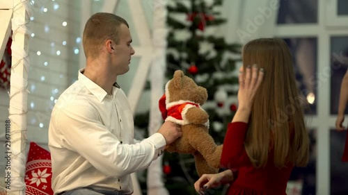 The family of mum, dad and daugher is kissing, hugging each other. The dad's giving a teddybear that wears a santahat to the small girl. There is  slightly blurred New Year background. photo