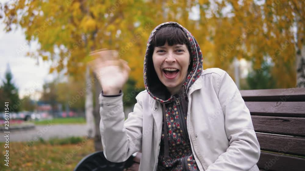 The girl sends a kiss looking at the camera. A young woman wearing a white jacket and hood sits on a bench against a background of yellow leaves. The girl sends a kiss looking at the camera. A young woman wearing a white jacket and hood sits on a bench against a background of yellow leaves.