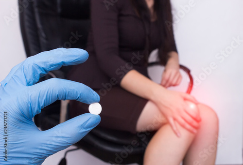 Business girl in the office sits in a computer chair and holds her knee, pain and inflammation in the knee, the doctor holds the pill in her hand