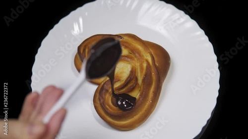 Someone pours chocolate on a sweet heart-shaped bun, fresh baking lies on a white plate and rotates counterclockwise on a black background, top view. photo