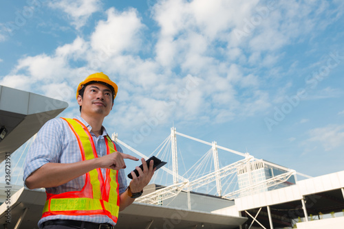 Young engineer wearing yellow safety helmet using a mobile phone in front construction building.