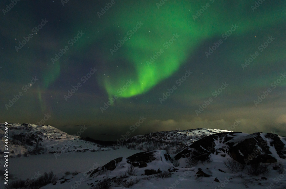 Beautiful northern lights, aurora in the night sky above the snow-covered hills. Large stones and a frozen lake.