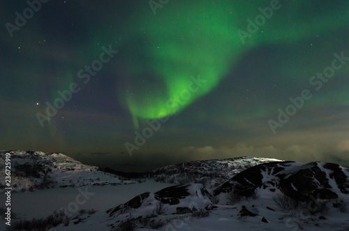 Beautiful northern lights, aurora in the night sky above the snow-covered hills. Large stones and a frozen lake. © Moroshka