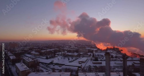 A large boiler room is located in the middle of a snowy city. From her five huge pipes, smoke and steam are pulled out of the air. It all sneaks up with a red sunshine.