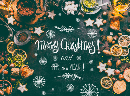 Merry Christmas greeting card with text lettering , spices, chocolate and cookies on dark background with sweet food ingredients: nuts, dried fruits, broken chocolate, cookies and winter spices