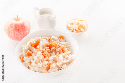 Oatmeal with pumpkin and nuts in a plate, an apple, sliced vegetables and a jug of milk on a white background. Close-up. Copy space