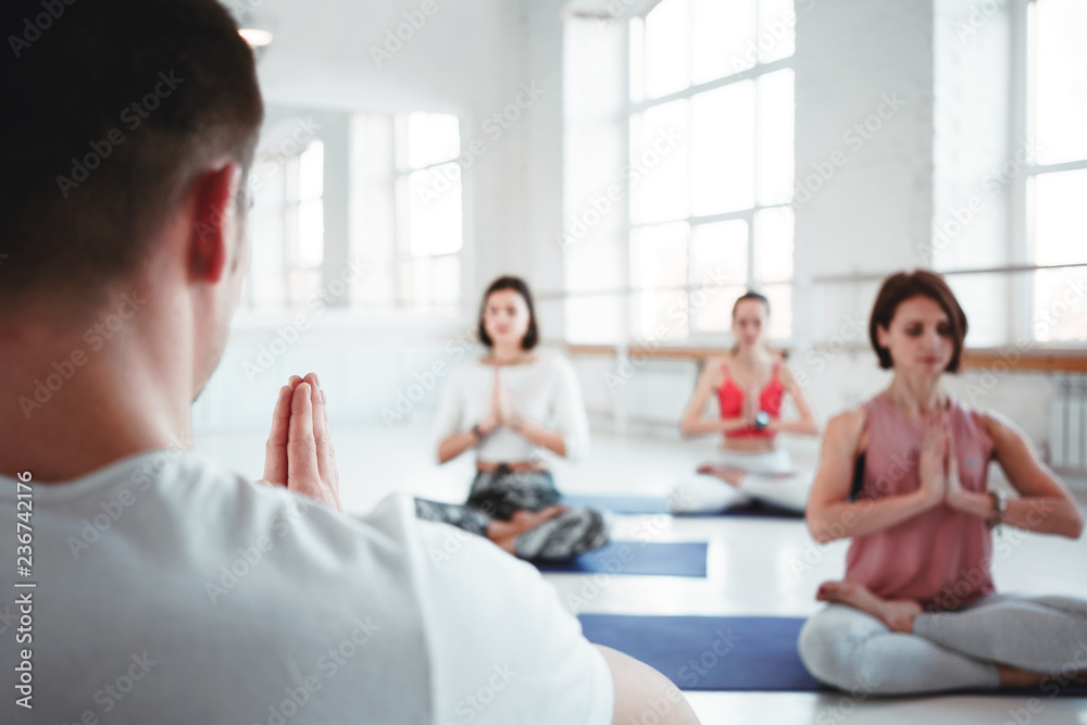 Fototapeta premium Man coach trains group of women yoga exercises for maintaining health care in white class. People together practis yoga poses. Blurred background