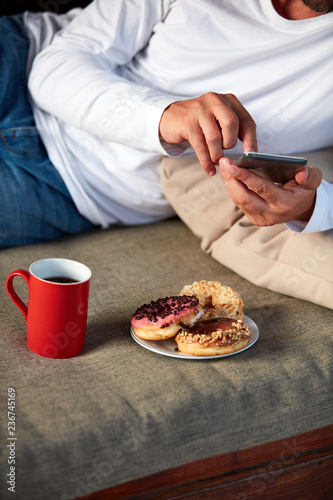 Man sitting on a terrace sofadrinking coffee and eating donuts while using cellphone.