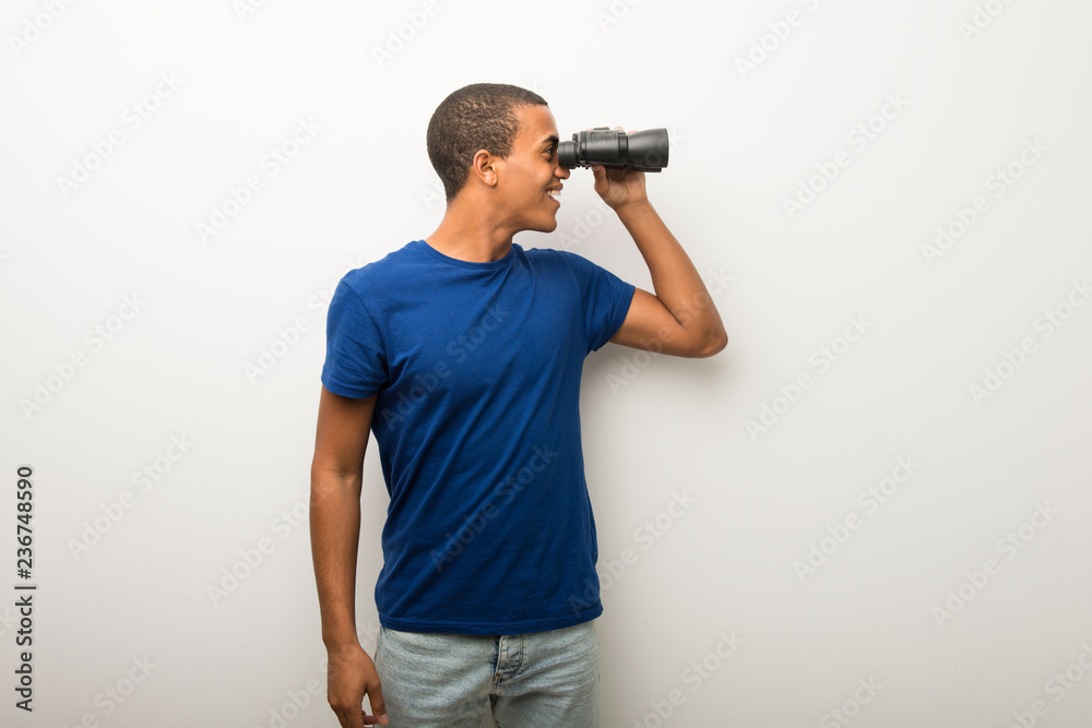 Young african american man on white wall and looking in the distance with binoculars Young african american man on white wall and looking in the distance with binoculars