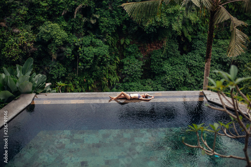 Slim sexy brunette woman in swimsuit relaxing on edge tropical infinity pool in jungle. Palms around and crystal clean water. Luxury resort on Bali island.