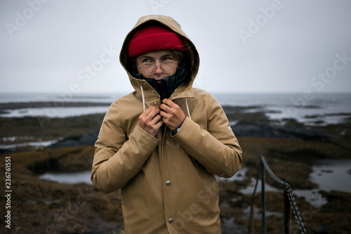 Cute and young woman shivers in cold rain and freezing wind on cloudy and gloomy day in iceland. Closes up beige raincoat to keep warmth inside. Concept winter tourism and adventures in scandinavia photo