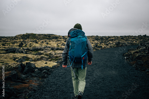 Back of traveler or hiker walking away into distance in waterproof jacket, beanie hat and backpack for camping gear. Epic grey and green moss covered icelandic landscape. adventure vibes lifestyle