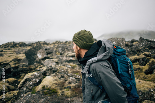 Young man in hiking trekking gear, waterproof jacket, green knit beanie and hike backpack walk through moss covered rough iceland terrain. Explore travel real wilderness lifestyle