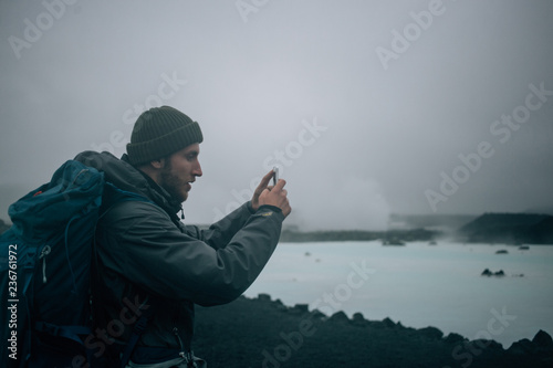 Young man in hiking outfit with camping bikepack makes photo on smartphone of icelandic landscape. Travel blogger adventure lifestyle. Winter hike in the wild photo