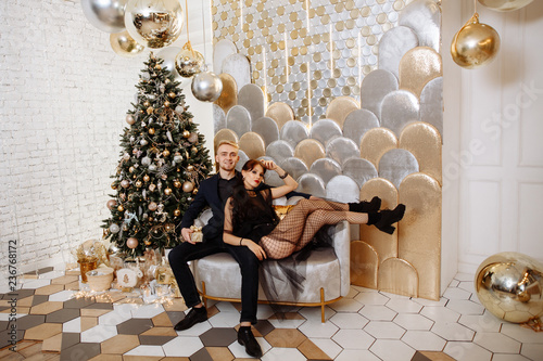 Christmas party concept. Stylishly dressed man and woman exchange gifts in a beautiful gold interior of the living room against the Christmas tree.