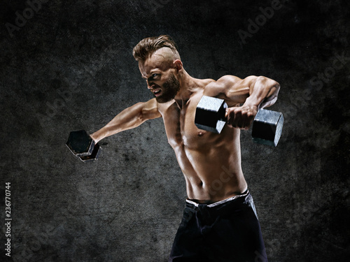 Bodybuilder doing the exercises with dumbbells. Photo of young man with naked torso on dark background. Strength and motivation.