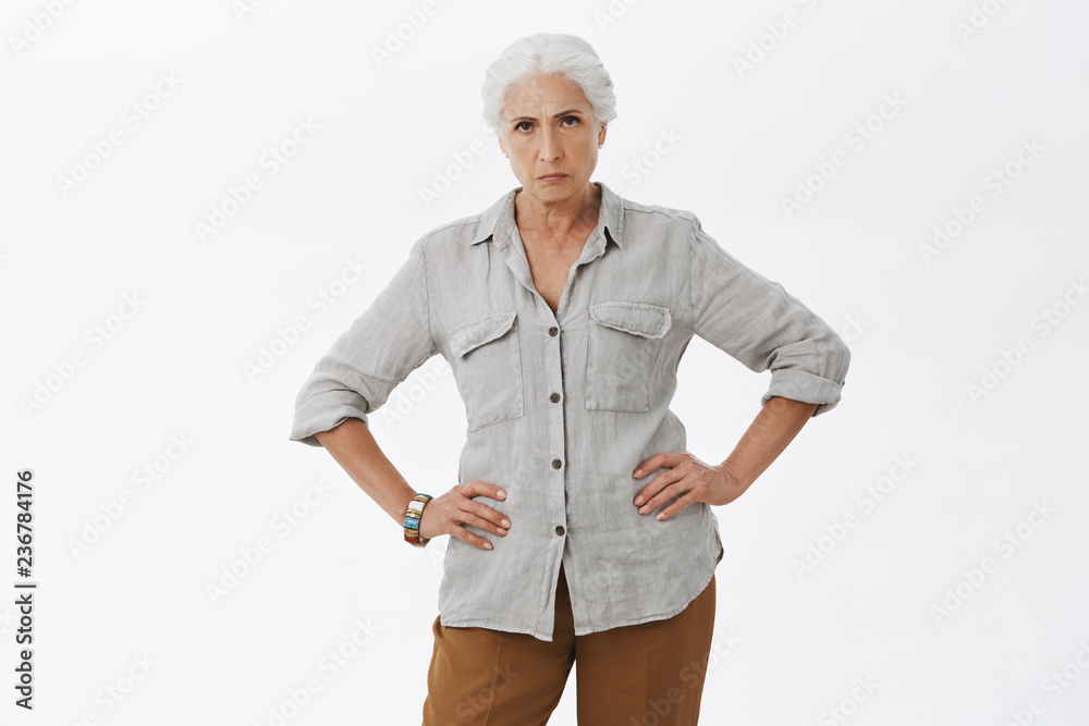 Explain you behavior young man. Portrait of strict and disappointed mad granny standing in displeased pose with hands in waist frowning staring angry at camera being dissatisfied posing over gray wall Explain you behavior young man. Portrait of strict and disappointed mad granny standing in displeased pose with hands in waist frowning staring angry at camera being dissatisfied posing over gray wall