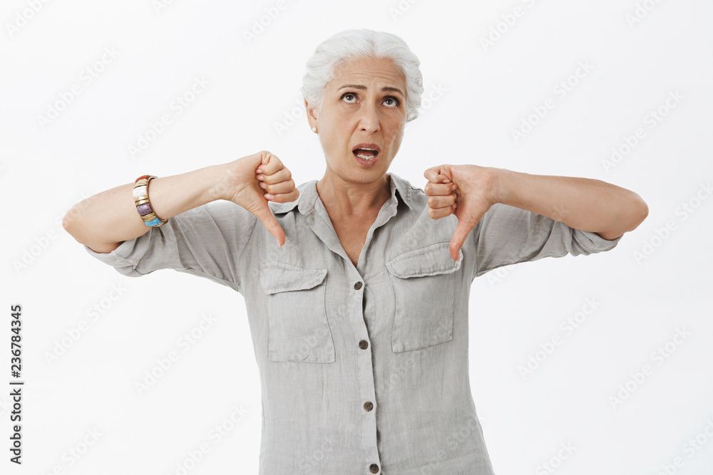 Even granny thinks it is lame. Energized unimpressed emotive elderly mother with grey hair in casual shirt rolling eyes up from boredom showing thumbs down in dislike gesture over grey wall Even granny thinks it is lame. Energized unimpressed emotive elderly mother with grey hair in casual shirt rolling eyes up from boredom showing thumbs down in dislike gesture over grey wall