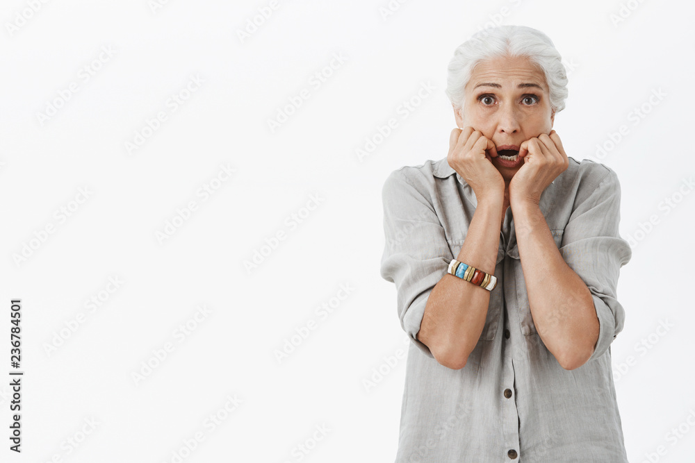 Waist-up shot of scared and concerned old lonely lady feeling unsafe and trembling from fear holding palms near mouth stooping standing worried and frightened against gray background
