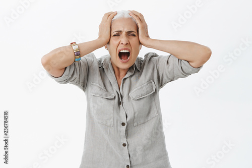 Studio shot of disstressed irritated and senior woman losing temper screaming out loud from pain and depression holding hands on head troubled and concerned being pissed and fed up photo